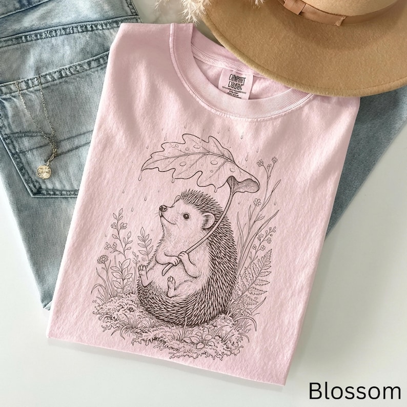 Puede incluir: Una sudadera Comfort Colors rosa claro con una ilustraci&oacute;n detallada de un erizo sosteniendo un paraguas de hojas. El erizo est&aacute; rodeado de plantas y flores. La palabra "Blossom" est&aacute; impresa en la esquina inferior derecha. Un sombrero color canela y unos vaqueros azules est&aacute;n en el fondo.