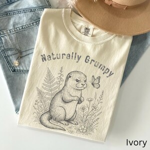 Puede incluir: Camiseta de color marfil con el texto "Naturally Grumpy" sobre una ilustración detallada de una nutria, una mariposa y elementos florales. La camiseta está hecha de un material suave y tiene un ajuste cómodo.