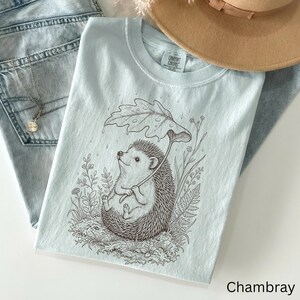 Puede incluir: Camiseta azul claro con un dibujo de un erizo sosteniendo un paraguas de hojas, rodeado de plantas. La palabra "Chambray" est&aacute; impresa en la esquina inferior derecha. La camiseta se muestra con unos vaqueros azules y un sombrero beige. La camiseta tiene el logo "Comfort Colors".