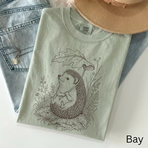 Puede incluir: Camiseta Comfort Colors verde claro con una ilustraci&oacute;n de un erizo bajo un paraguas de hojas. La camiseta se muestra con vaqueros azul claro y un collar. La palabra "Bay" est&aacute; impresa en la parte inferior derecha.