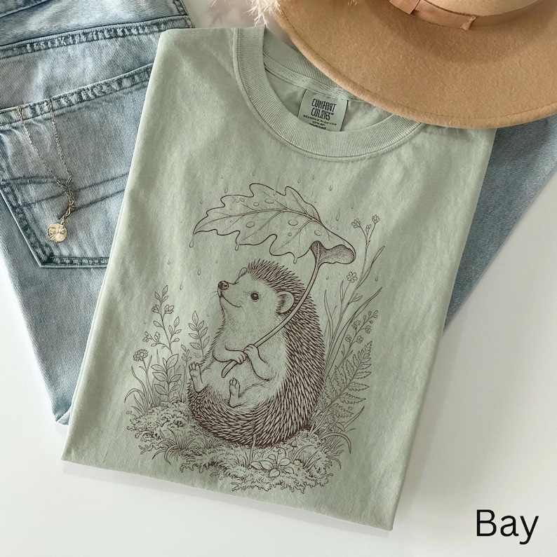 Puede incluir: Camiseta Comfort Colors verde claro con una ilustraci&oacute;n de un erizo bajo un paraguas de hojas. La camiseta se muestra con vaqueros azul claro y un collar. La palabra "Bay" est&aacute; impresa en la parte inferior derecha.