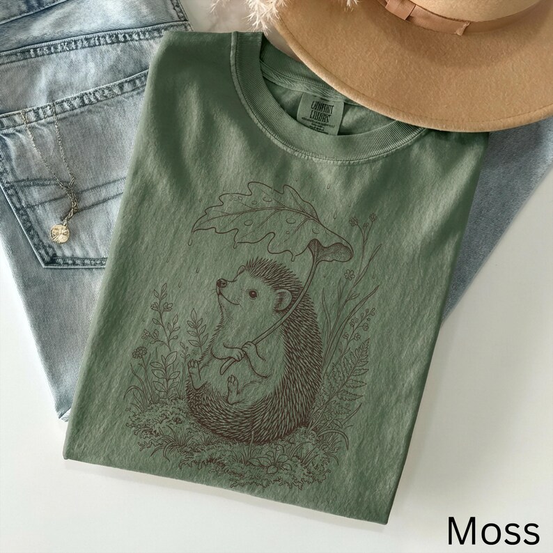 Puede incluir: Camiseta verde musgo con una ilustraci&oacute;n marr&oacute;n de un erizo bajo una hoja. La camiseta se combina con vaqueros azul claro y un sombrero beige. La palabra "Moss" est&aacute; impresa en la parte inferior. La camiseta es de material suave.
