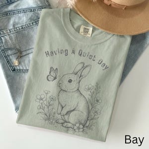 May include: A light green t-shirt with the text "Having A Quiet Day" and a graphic of a rabbit, butterfly, and flowers. The shirt is laid on top of blue jeans and a tan hat.