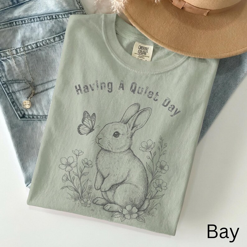 May include: A light green t-shirt with the text "Having A Quiet Day" and a graphic of a rabbit, butterfly, and flowers. The shirt is laid on top of blue jeans and a tan hat.