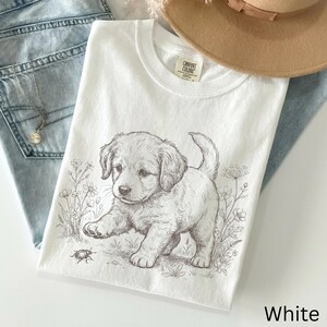 May include: White t-shirt with a detailed illustration of a puppy and a beetle amongst flowers. The shirt is made of a soft material and is labelled "Comfort Colors". The word "White" is printed on the bottom right.