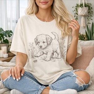 May include: Cream-coloured t-shirt with a detailed sketch of a golden retriever puppy exploring a field of flowers and a small insect. The graphic is in black and white. The shirt is paired with blue jeans.
