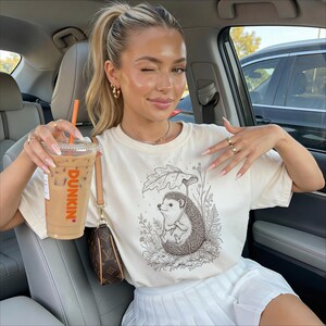 Puede incluir: Camiseta color crema con una ilustraci&oacute;n detallada de un erizo sosteniendo un paraguas de hojas. La camiseta se combina con una falda plisada blanca. Una taza transparente de caf&eacute; helado con el logo de Dunkin' se sostiene en la mano.
