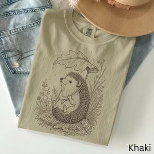 Puede incluir: Camiseta color caqui con una ilustraci&oacute;n detallada de un erizo sosteniendo un paraguas de hoja, rodeado de plantas. La camiseta tiene cuello redondo y mangas cortas. La palabra "Khaki" est&aacute; impresa en la parte inferior derecha.