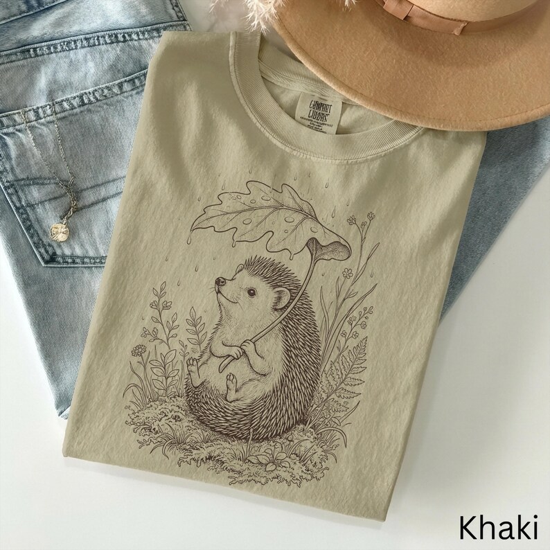 Puede incluir: Camiseta color caqui con una ilustraci&oacute;n detallada de un erizo sosteniendo un paraguas de hoja, rodeado de plantas. La camiseta tiene cuello redondo y mangas cortas. La palabra "Khaki" est&aacute; impresa en la parte inferior derecha.