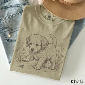 May include: Khaki t-shirt with a detailed sketch of a puppy and a bug, surrounded by floral elements. The shirt is folded on blue jeans, with a necklace and a tan hat visible. The word "Khaki" is at the bottom.