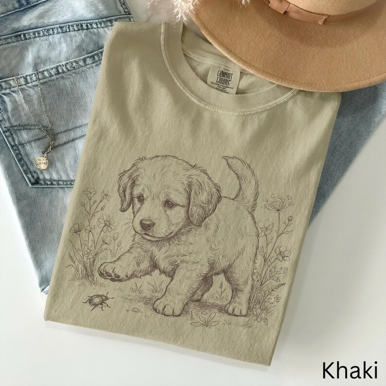 May include: Khaki t-shirt with a detailed sketch of a puppy and a bug, surrounded by floral elements. The shirt is folded on blue jeans, with a necklace and a tan hat visible. The word "Khaki" is at the bottom.