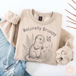 Könnte beinhalten: Sandfarbenes Sweatshirt mit dem Text "Naturally Grumpy" über einer detaillierten Illustration eines Otters, eines Schmetterlings und floralen Elementen. Das Sweatshirt hat einen Rundhalsausschnitt und ist aus weichem Material.