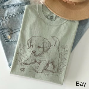 May include: A pale green Comfort Colors t-shirt with a detailed illustration of a puppy and a bug. The puppy is light brown, and the shirt has a crew neck. The word "Bay" is printed in the bottom right corner.