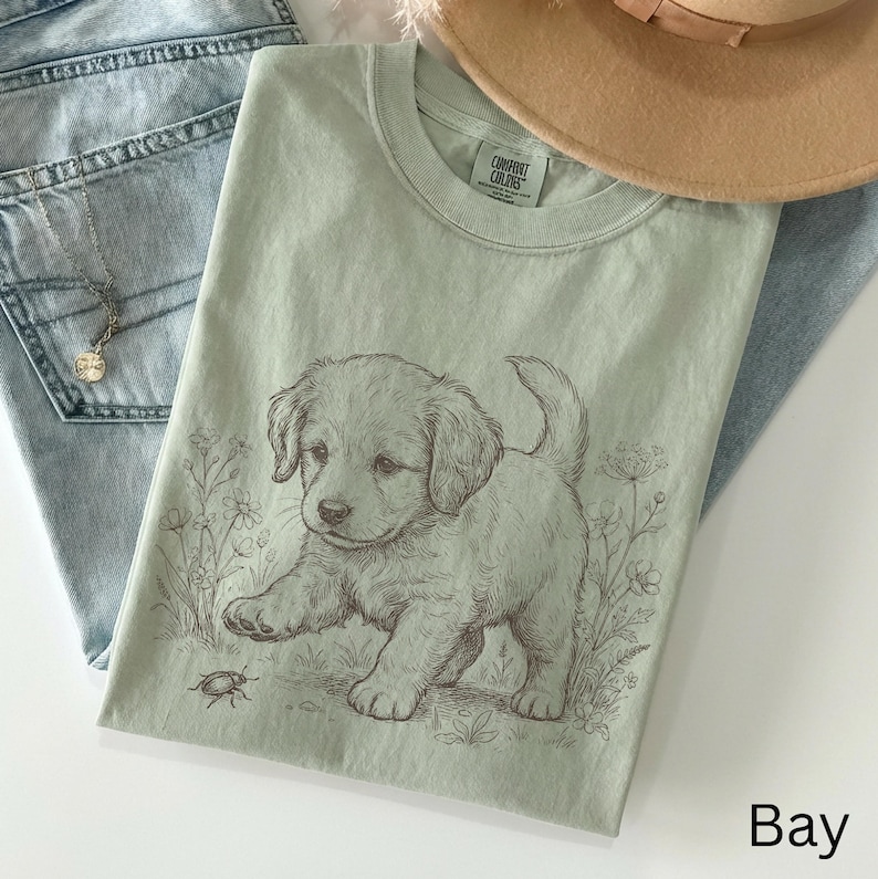 May include: A pale green Comfort Colors t-shirt with a detailed illustration of a puppy and a bug. The puppy is light brown, and the shirt has a crew neck. The word "Bay" is printed in the bottom right corner.