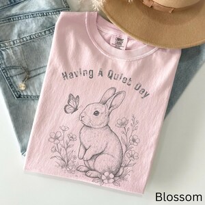 May include: A light pink t-shirt with the text "Having A Quiet Day" and a graphic of a rabbit and butterfly. The shirt is folded on top of blue jeans and a tan hat.
