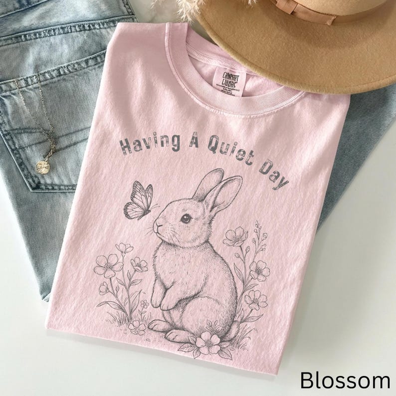 May include: A light pink t-shirt with the text "Having A Quiet Day" and a graphic of a rabbit and butterfly. The shirt is folded on top of blue jeans and a tan hat.