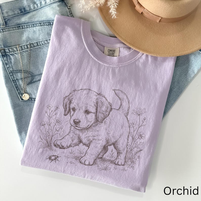 May include: A lilac-coloured t-shirt with a detailed illustration of a puppy playing with a bug in a field of flowers. The shirt is made of soft material and is folded on top of a pair of blue jeans. A gold necklace and a tan hat are also visible.