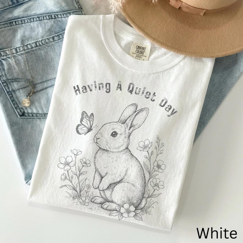 May include: White t-shirt with a graphic of a rabbit, butterfly, and flowers, with the text "Having A Quiet Day". The shirt is folded and placed on top of blue jeans and a tan hat.
