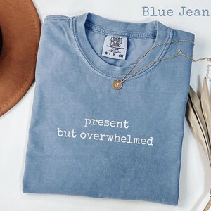 Puede incluir: Una camiseta azul vaquero con las palabras "present but overwhelmed" en blanco. Un collar dorado con un colgante está drapeado sobre la camisa. Un sombrero marrón está en la esquina superior izquierda.