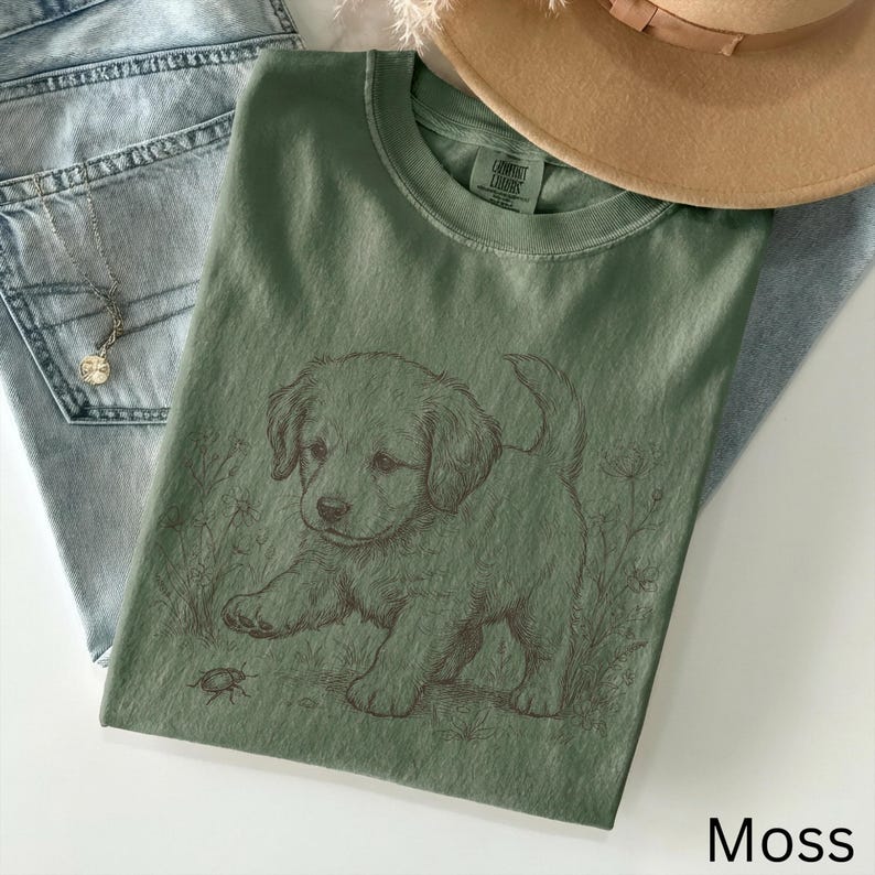 May include: A moss green t-shirt with a detailed illustration of a puppy and a bug. The shirt is made of a soft material and is paired with light blue jeans and a tan hat. The word "Moss" is printed at the bottom.
