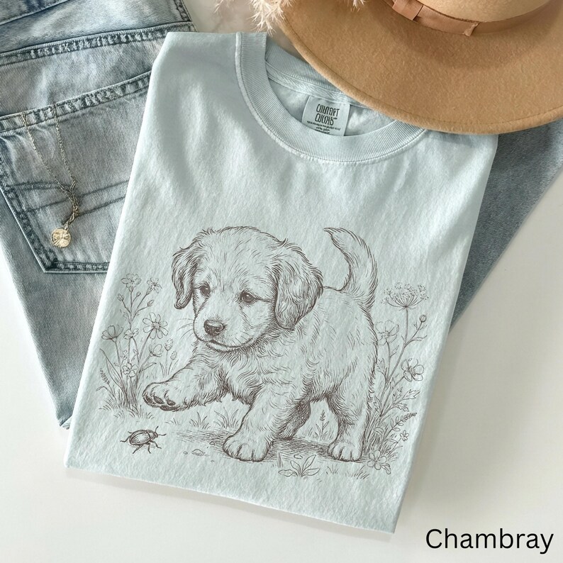 May include: Pale blue t-shirt featuring a puppy illustration reaching for a bug. The shirt has a crew neck and short sleeves. The text "Chambray" is at the bottom right. The shirt is on light blue jeans.