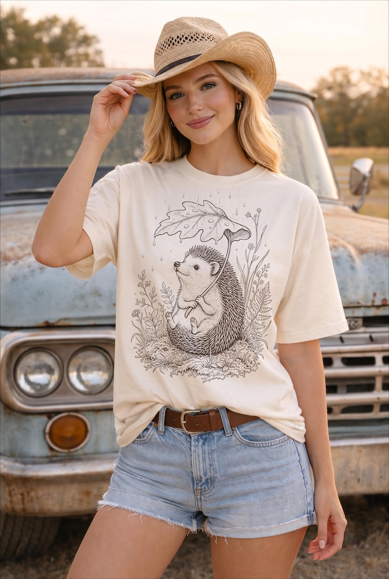 Puede incluir: Camiseta color crema con una ilustraci&oacute;n en blanco y negro de un erizo bajo un paraguas de hojas. Se combina con pantalones cortos de mezclilla y un cintur&oacute;n marr&oacute;n. Tambi&eacute;n se usa un sombrero de vaquero de paja.