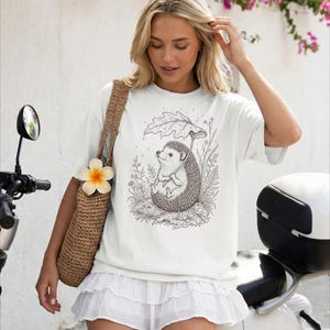 Puede incluir: Camiseta blanca con una ilustraci&oacute;n en blanco y negro de un erizo sosteniendo un paraguas de hoja bajo la lluvia. El erizo est&aacute; rodeado de plantas. La camiseta la lleva una persona.