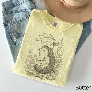 Puede incluir: Camiseta amarillo claro con un dibujo de un erizo bajo una hoja. La camiseta est&aacute; colocada sobre unos vaqueros azules, un collar y un sombrero beige. La palabra "Butter" est&aacute; impresa en la parte inferior derecha.