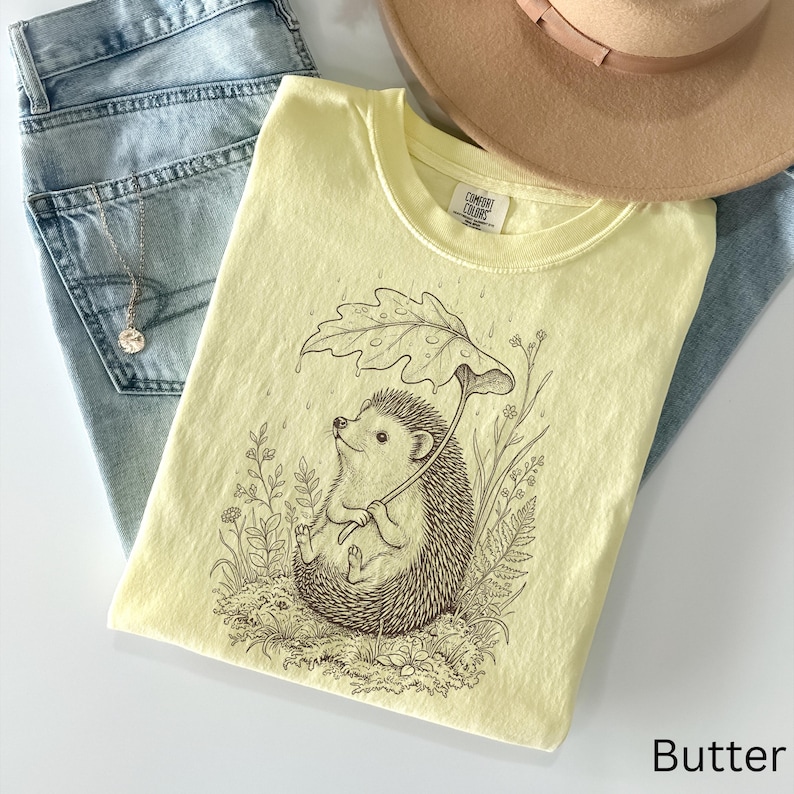 Puede incluir: Camiseta amarillo claro con un dibujo de un erizo bajo una hoja. La camiseta est&aacute; colocada sobre unos vaqueros azules, un collar y un sombrero beige. La palabra "Butter" est&aacute; impresa en la parte inferior derecha.