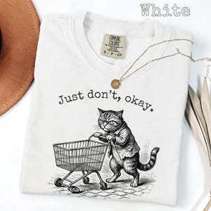 May include: White t-shirt with the text "Just don't, okay." and a graphic of a cat pushing a shopping cart. The cat is wearing a suit jacket. A gold necklace is draped across the shirt.