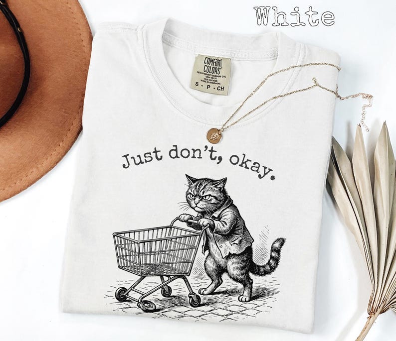 May include: White t-shirt with the text "Just don't, okay." and a graphic of a cat pushing a shopping cart. The cat is wearing a suit jacket. A gold necklace is draped across the shirt.