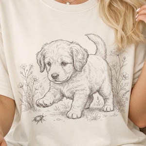 May include: A cream-coloured t-shirt with a black and white illustration of a golden retriever puppy in a field of flowers. The puppy is looking at an insect. The shirt has a relaxed fit.