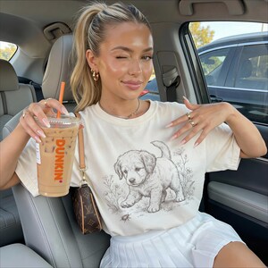 May include: A cream-coloured t-shirt with a detailed sketch of a puppy. The shirt is worn with a white pleated skirt. A clear iced coffee cup with the Dunkin' logo is held. The person is sitting in a car. The image has a summery feel.