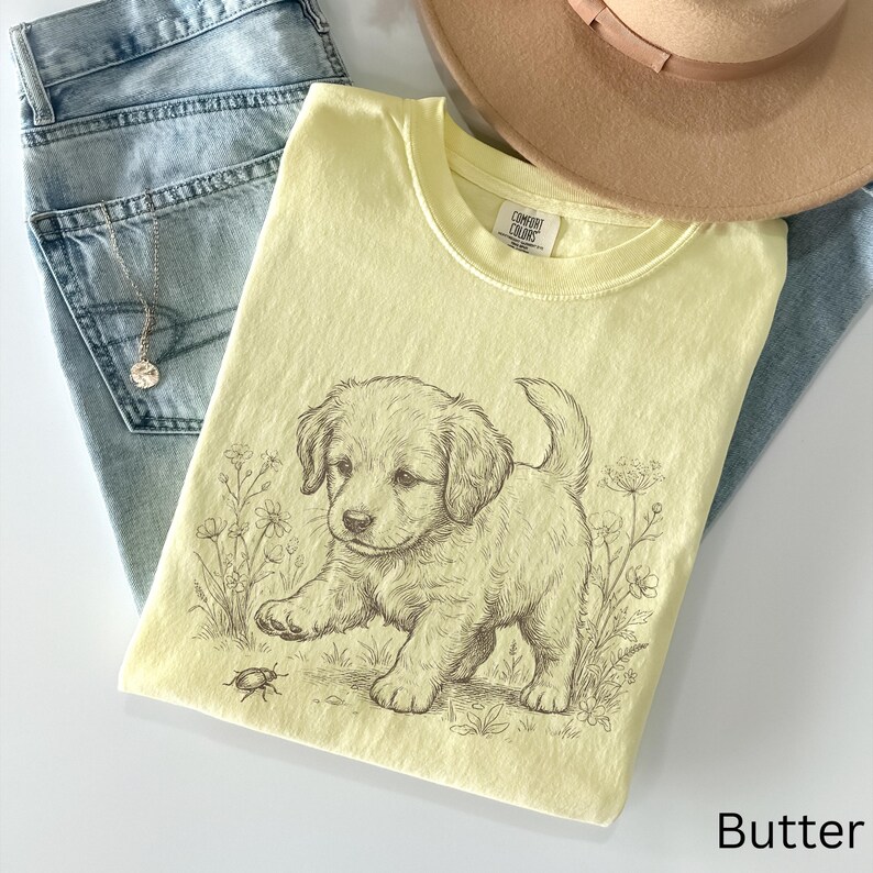 May include: A pale yellow t-shirt featuring a puppy illustration, laid on blue denim jeans and a beige hat. The t-shirt has the word "Butter" printed at the bottom. The shirt is folded.