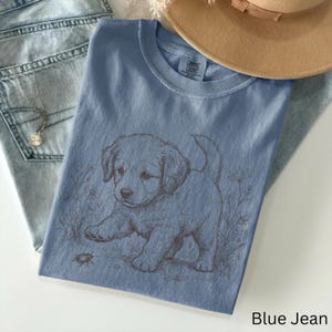May include: A blue t-shirt featuring a puppy graphic and the text "Blue Jean". The puppy is light brown and is reaching towards a small insect. The shirt is next to a pair of blue jeans and a tan hat.