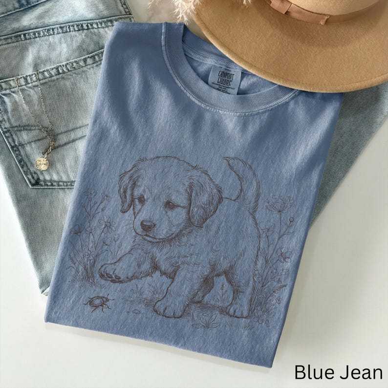 May include: A blue t-shirt featuring a puppy graphic and the text "Blue Jean". The puppy is light brown and is reaching towards a small insect. The shirt is next to a pair of blue jeans and a tan hat.