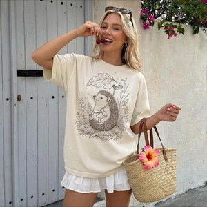 Puede incluir: Camiseta color crema con una ilustraci&oacute;n detallada de un erizo bajo un paraguas de hojas. La camiseta se combina con una falda blanca y un bolso tote tejido con una flor rosa y amarilla. La persona sostiene cerezas.