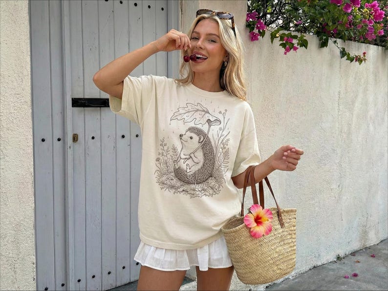 Puede incluir: Camiseta color crema con una ilustraci&oacute;n detallada de un erizo bajo un paraguas de hojas. La camiseta se combina con una falda blanca y un bolso tote tejido con una flor rosa y amarilla. La persona sostiene cerezas.