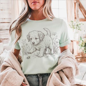May include: A pale green t-shirt with a detailed sketch of a puppy walking through a field of flowers. The puppy is the focal point, rendered in black and white, with a small insect in front of it. The shirt has rolled-up sleeves and is paired with blue jeans.