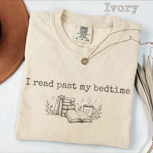 May include: Ivory-colored t-shirt with the text "I read past my bedtime" and a graphic of books, a mug, and floral accents. A gold necklace with a round pendant is draped on the shirt. The shirt is folded on a white surface.