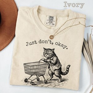 May include: Ivory-colored t-shirt with the text "Just don't, okay." and a graphic of a cat pushing a shopping cart. The cat is wearing a suit. A gold necklace with a pendant is on the shirt.