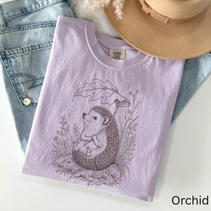 Puede incluir: Una camiseta de color lila claro con un dibujo de un erizo sosteniendo un paraguas de hojas, rodeado de plantas. La camiseta est&aacute; etiquetada como "Comfort Colors" y el color es "Orchid". La camiseta est&aacute; doblada sobre unos vaqueros.