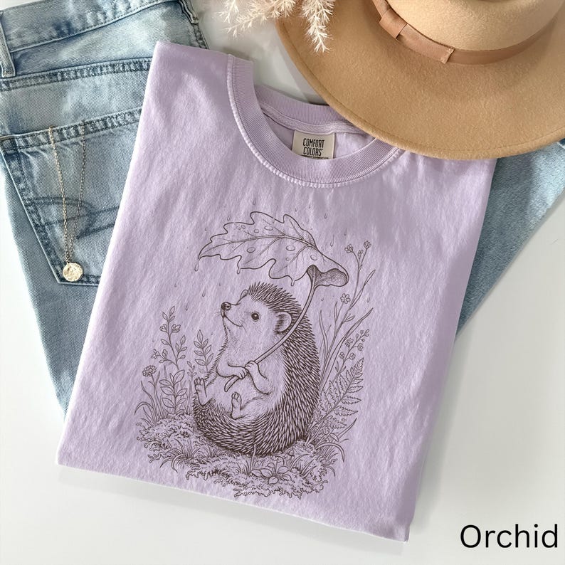 Puede incluir: Una camiseta de color lila claro con un dibujo de un erizo sosteniendo un paraguas de hojas, rodeado de plantas. La camiseta est&aacute; etiquetada como "Comfort Colors" y el color es "Orchid". La camiseta est&aacute; doblada sobre unos vaqueros.