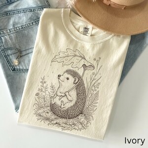 Puede incluir: Camiseta de color marfil con una ilustraci&oacute;n caprichosa de un erizo sosteniendo un paraguas de hojas. El dise&ntilde;o est&aacute; en un estilo de arte lineal detallado, con el texto "Comfort Colors" visible en la etiqueta. La camiseta se combina con vaqueros azules y un sombrero marr&oacute;n.