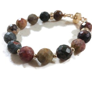 May include: A bracelet made of faceted gemstone beads in shades of brown, green, and red. The beads are connected with gold-toned metal spacers and a magnetic clasp.