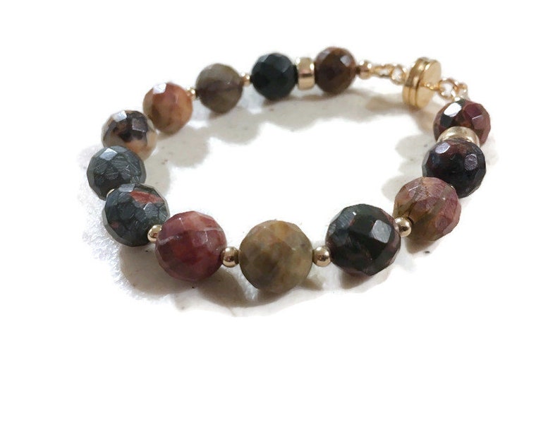 May include: A bracelet made of faceted gemstone beads in shades of brown, green, and red. The beads are connected with gold-toned metal spacers and a magnetic clasp.