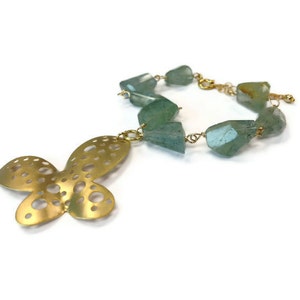 Puede incluir: Un dije de mariposa dorado cuelga de un collar hecho de cuentas de piedras preciosas verde claro y una cadena de oro.