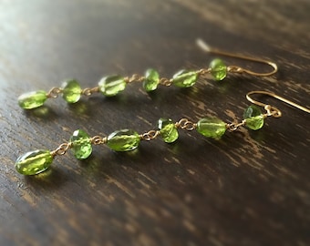 May include: A pair of long, green gemstone earrings with gold wire and hooks. The earrings are made with oval-shaped green stones and gold wire.
