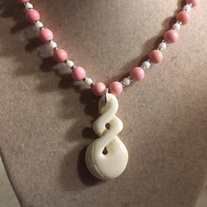 May include: A white carved pendant necklace with a pink and white bead chain. The pendant is a twisted rope design.