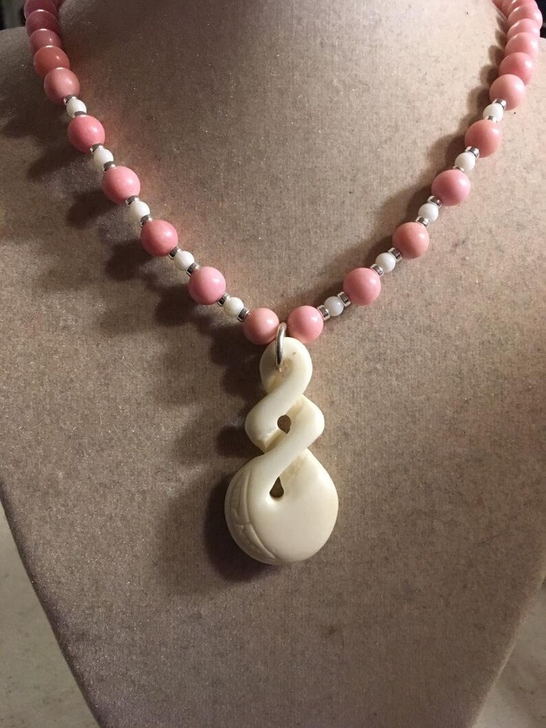 May include: A white carved pendant necklace with a pink and white bead chain. The pendant is a twisted rope design.